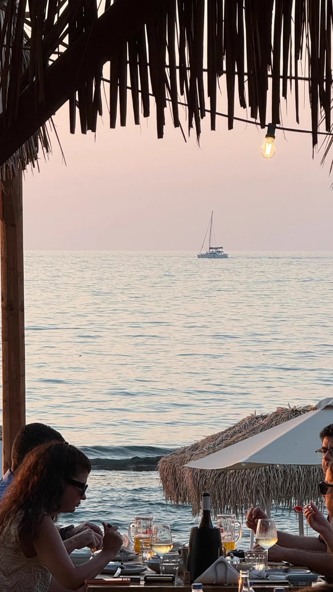 Sunset dining under a reed roof with wine glasses and a sailboat on the horizon