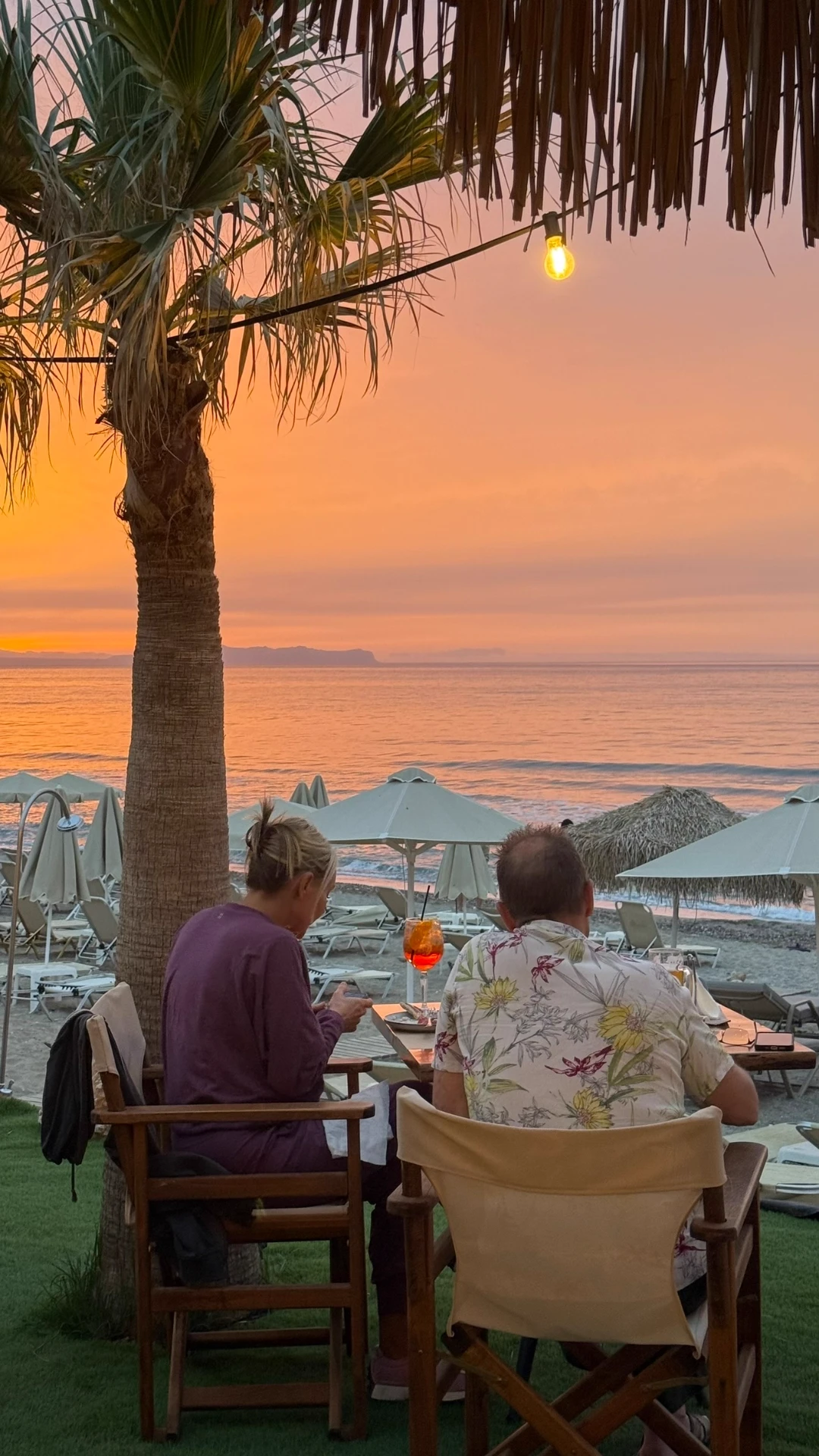 Couple enjoying a cocktail at a beachfront table under a palm tree at sunset