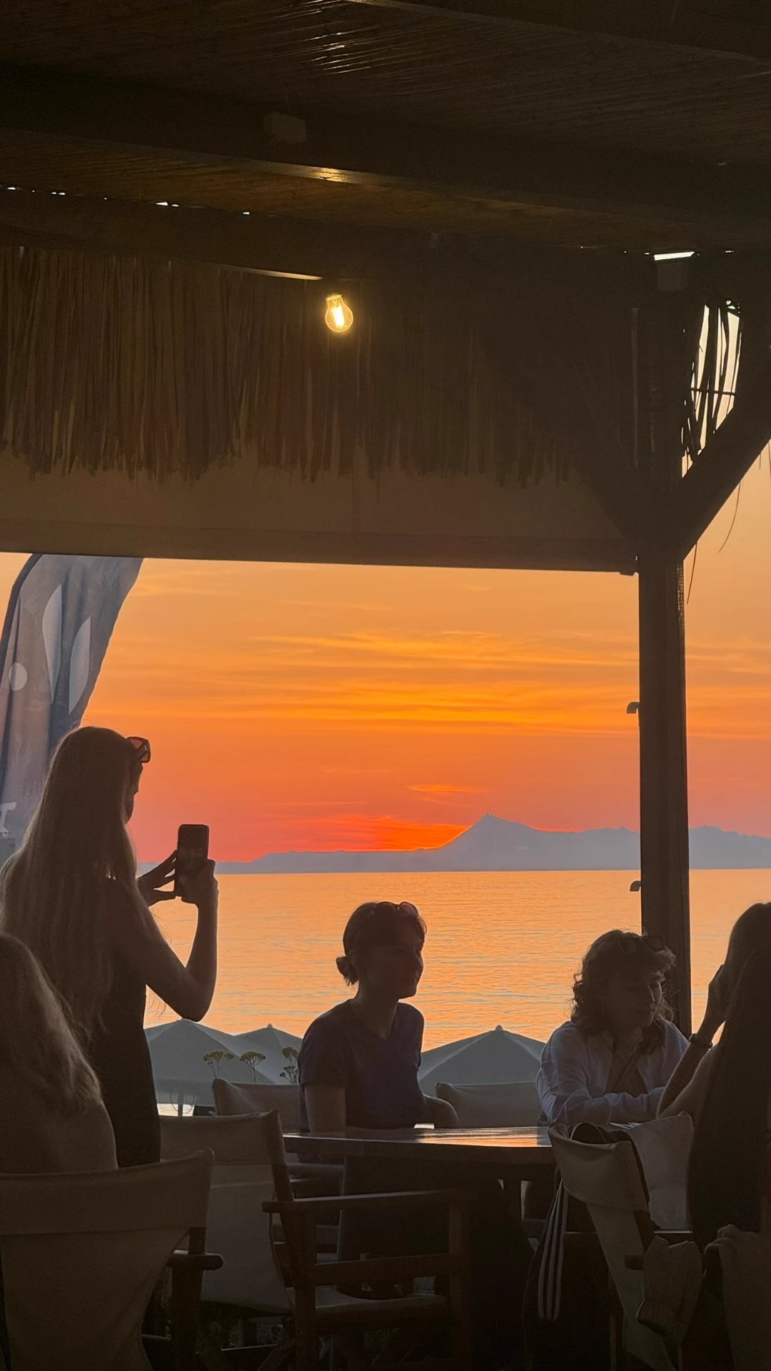 Friends gathered around a table under the reed roof, silhouetted against a deep orange sunset