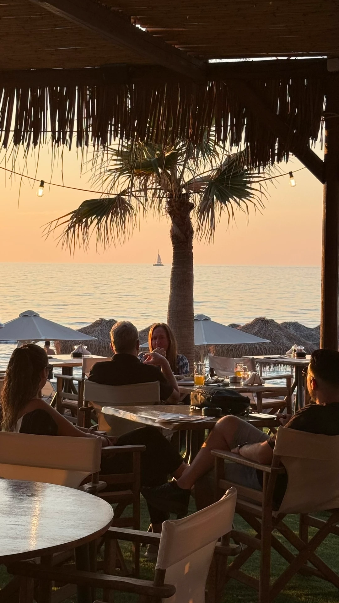 Guests dining on the beachfront terrace at sunset with a sailboat passing